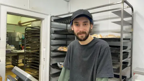 BBC/Emily Johnson A man with a beard, hairnet and hat stands at the till in the bakery.