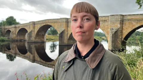 BBC Sophia Stovell standing in front of the Chollerford Bridge, a long stone structure with five arches, over the River Tyne. She is standing on the river bank at the George Hotel in Chollerford. She is wearing a brown and green jacket with a black shirt. Her hair is light brown and has a short fringe. 