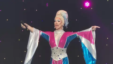 Peterborough Pride Divina de Campo is on a stage. They are wearing an elaborate pink, white and blue outfit and have hair the same colours.