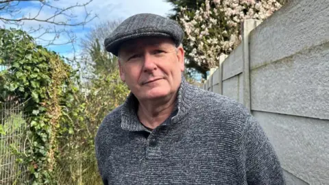 BBC/ Alex James A man is stood in an alley next to a white fence. He is wearing a grey flat cap and a grey jumper. To the left of the image is a chain link fence covered in ivy. 