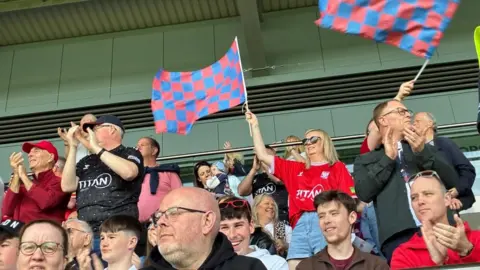 A group of York City FC football fans sit in the stands, waving large blue and red checked flags.
