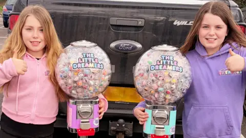 Little Dreamers Two girls in pink and purple hoodies with thumbs up, beside two of their spherical vending machines