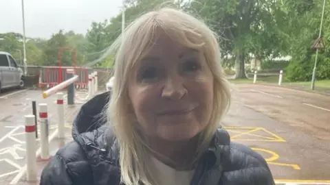 Piers Hopkirk/BBC A blonde woman smiling at the cameras. She is wearing a black coat and there is a car park behind her.