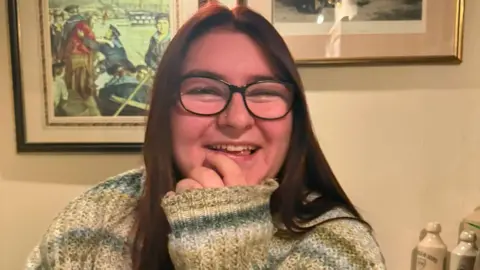 Bethany Lewis A young woman with brown/red hair and black glasses is smiling. She has a stud on her nose and is wearing a green, blue and cream jumper with her hand to her mouth