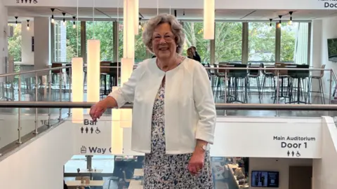 Fareham Live Councillor Connie Hockley  standing in the theatre foyer wearing a flowery dress and white jacket, smiling at the camera she has glasses on.