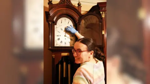 Picture of a young brown haired woman wearing glasses in a rainbow striped jumper. She is changing the time on a grandfather clock, wearing blue gloves and is smiling at the camera.