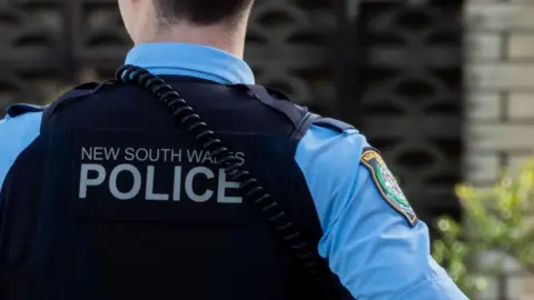 The back profile of a police officer's uniform. The officer wears a blue shirt and navy vest with 'New South Wales Police' printed on the back in white. A radio wire drapes over their shoulder. The right hand sleeve of the shirt has a navy, yellow and green badge on it. In the background is a blurred residential building.