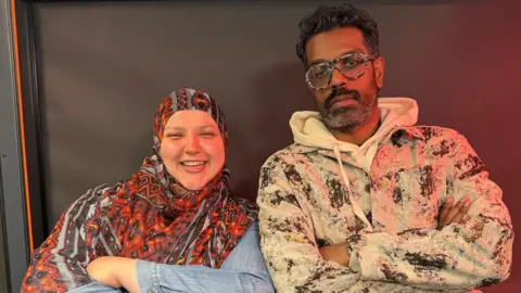 A woman wearing an orange, patterned headscarf smiles at the camera. She stands next to a man with short, curly hair wearing glasses. He looks at the camera. They both have their arms folded.