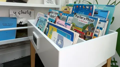 A white box with three rows of colourful children's books in Manx lined up. There are more on another white shelf to the left.