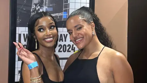 Vanessa Marie, a female, on the right with a female friend on the left in front of a poster for the Youth Music awards on a light brown wall. They are both wearing black tops and smiling. 