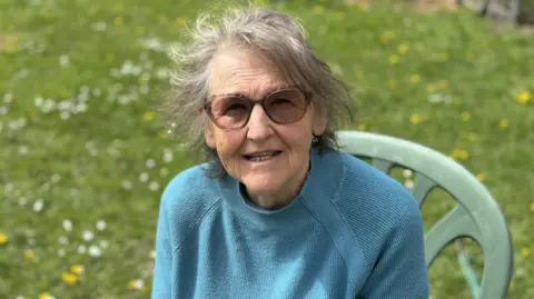 Luke Deal/BBC Sandy Walne sits in a chair outside on a grassed area. She has short grey hair and wears sunglasses with a blue jumper.