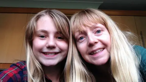 A young girl smiles in a selfie with her mother, in front of kitchen cupboards. Both have blonde hair.