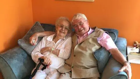 Bernard O’Malley (right) has his arm around his wife, Joan. The pair are both smiling and sitting on a grey double seater sofa  