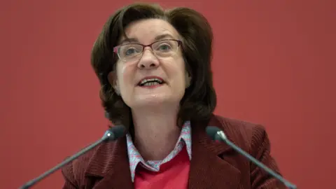 Eluned Morgan is stood looking up from a lecturn which has microphones on top of it. She is wearing a dark red jacket and a red top with a shirt underneath.