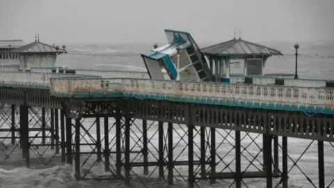 An overturned bluish  and achromatic  hut leans against different  hut connected  Llandudno Pier, with ample  waves disposable   each  around. Two different   huts tin  beryllium  seen further on  the pier. 