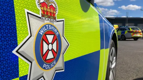 BBC The side of a marked Wiltshire Police car, which is parked in a car park. 