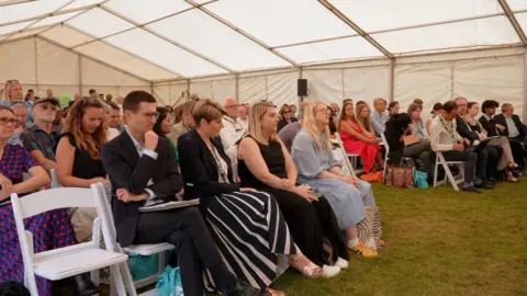 Yvette Austin/BBC Visitors inside a marquee at the River Summit hear about river protection from various speakers. They are sitting on white chairs on grass.
