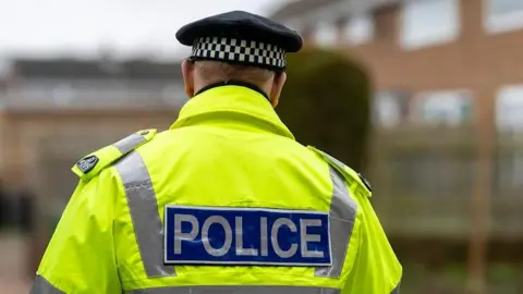 Police officer wearing a hi-vis jacket with the word: "Police" written on the back. He is walking around what looks like a housing estate.