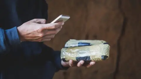 man's hands shown carrying a suspicious-looking packet wrapped in masking tape and a mobile phone