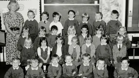A black and white photo of Gwen Mayor, a blonde-haired woman in a patterned dress, and her 28 pupils arranged in four rows of seven in a classroom. The children are in school uniform.