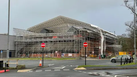 Metal scaffolding surrounding a brick building. White sheets have fallen off the roof.