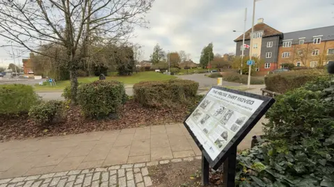 Steve Hubbard/BBC A black information board featuring information and pictures on a white board stands at waist height next to a paved walkway which runs next to a roundabout, which is seen in the background alongside some flats.