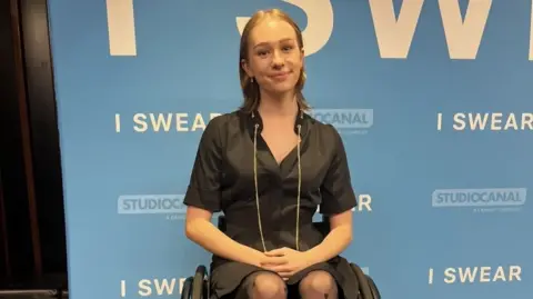 A woman in a wheelchair wearing a black dress in front of a promotional board for the film I Swear