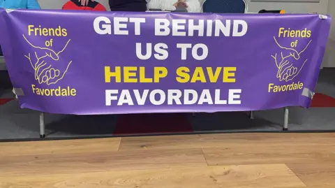 A purple sign with the words "Get behind us to help save Favordale" in white and yellow lettering.  The banner has the Friends of Favordale logo on either side.