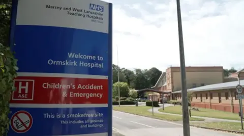 Close up of a sign which reads 'Welcome to Ormskirk Hospital' at the entry to the hospital building in the background on a clear day.