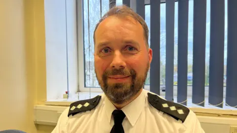 Ellen Knight/BBC CI Parker is photographed looking into the camera and smiling slightly. He is wearing a white collared shirt with a black tie, and has black epaulettes with three white diamond shapes on them, indicating his rank. He has short light brown hair, and a short brown beard. He is sat down in a room with white walls, in front of a window with a blue blind. The sky outside the window is blue with clouds. 