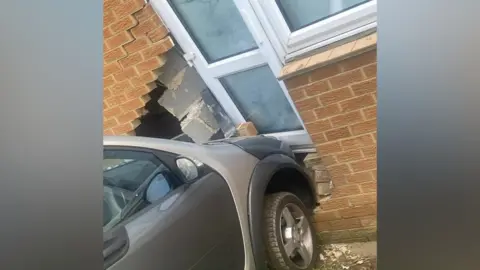 Chloe Beale A small grey car has smashed a hole in the brick exterior of a house and pushed the white patio door off its hinges. There is debris scattered around from the damage. A section of brickwork is missing, behind which are broken building blocks and a view into the damaged living room.