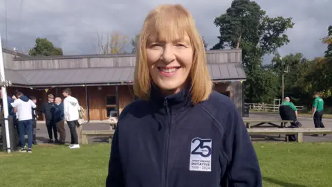 Kate Thompson is smiling into the camera. She has shoulder-length fair hair. She's wearing a navy hoodie that commemorates 25 years of the joint award initiative. She's standing in a field and there are some young people in the background.