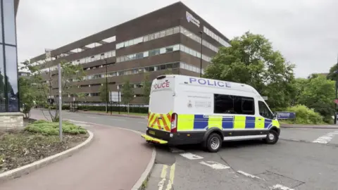 A large police van is parted across the road on the edge of a roundabout. Behind it is a large brown brick building with windows along each floor. Trees fringe the road ahead. To the left is the corner of an entirely glass building, the glass appearing black and reflecting the surroundings. On the ground in front of it is some scrubby soil.