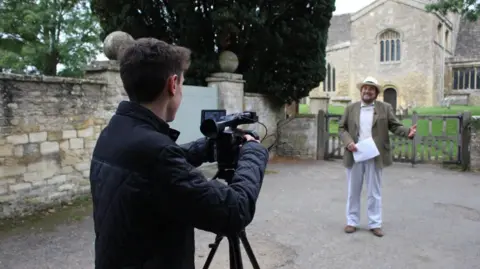 The Cotswold Explorer Robin Shuckburgh and his producer Ross Arrowsmith filming in front of a Cotswold church. Mr Shuckburgh is smiling while holding a piece of paper. Mr Arrowsmith is behind the camera filming him. It is a cloudy day.