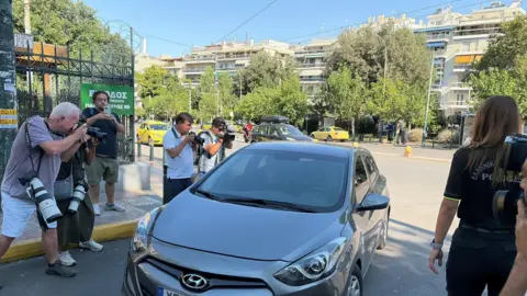 PA Media Several photographers standing on a city street photograph a silver Hyundai car parked at the roadside. In the foreground, camera operators with large telephoto lenses aim toward the vehicle, while a person in a dark uniform stands nearby. Trees, apartment buildings, yellow taxis, and Greek-language street signage are visible in the background under clear daytime skies.