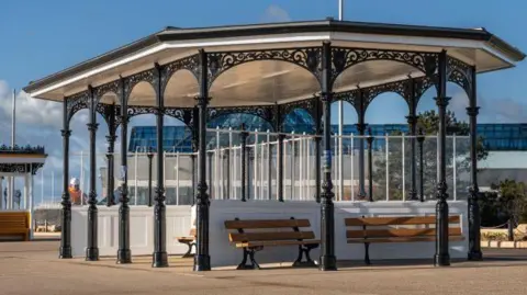 A large Victorian seafront shelter, resembling a large gazebo, is situated on the seafront. The columns are painted jet black, with wooden benches and white walls in the interior of the shelter. It's a sunny day and the shelter is well-lit. 