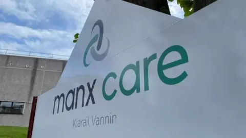 "Manx Care in green and black text on a white sign with a celtic knot on a sign outside a hospital building. 