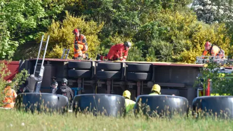 Pacemaker the underside of a lorry can be seen after it overturned. there are a number of rescue workers on top of the lorry and standing to its side. The area is surrounded in green bushes, trees and long grass.