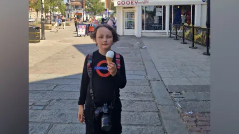 Supplied A picture of George stood outside a 'Gooey Waffle' shop in London on the day of the challenge. George is wearing all black, and his top has a TfL roundel on it. He has longish brown hair and is holding an ice cream. He also has a bag on his back and a camera around his neck.