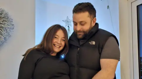 NHSBT Maisie, with husband Keith, stand indoors facing the camera, with a window, indoor plants and decorative star visible in the background.