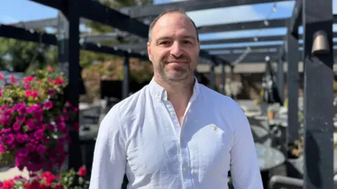 Photo of a man with short hair and stubble looking into the camera. He is wearing a white button-down shirt.