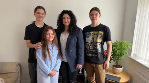 Nataliya Korovina is standing with her three children. She has dark long curly hair and is wearing a navy suit jacket and white shirt. Her two sons stand on either side of her, they both have long brown hair tied back in ponytails. In front of them is Nataliya's daughter, who has very long light brown hair and is wearing a blue shirt.