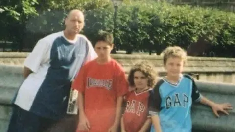 Family Handout An old image of Chris Coyle with his three eldest sons, It's a sunny day and the boys are young. Chris is tall with a bald head, two boys on the right of the picture, and to Chris's left, are wearing red t-shirts, while on the end is a boy in a light blue t-shirt.