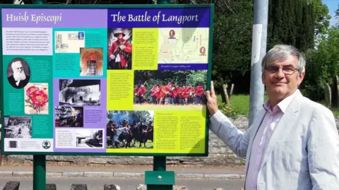 Julian Humphrys Julian Humphrys outside Huish Epsicopi next to a notice board reading 'The Battle of Langport'. He is wearing a grey blazer with a pink shirt. He has grey hair and glasses.