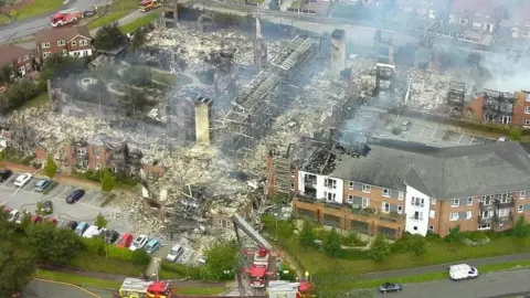 Cheshire Fire and Rescue Service An aerial view of a large burnt out building, its carcass still smouldering. There are fire crews with fire engines at various points around the structure.