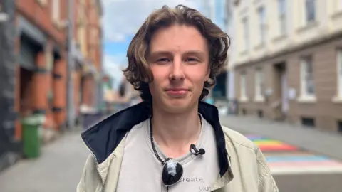 Max looks into the camera in front of a colourful street in Nottingham city centre. He is wearing a jacket and has his sunglasses hooked on the neck of his t-shirt.