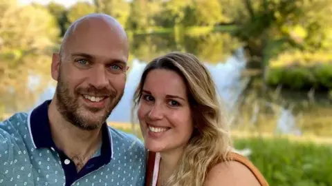 Ainara Pederoso A couple smile at the camera. A man with a light beard wearing a light blue patterned polo shirt is to the left of the picture. A woman with shoulder length blonde hair stands beside him.