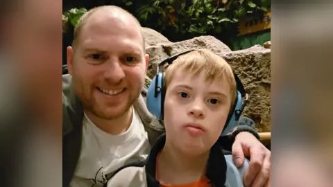 Colin Jones Colin and Coby pose for a selfie. Colin smiles with arm arm around Coby's shoulder. Both are wearing fleeces and t-shirts and Coby is wearing blue ear defenders.