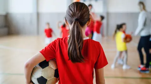 Rear view of small girl standing with ball indoors in gym class, physical education concept. 