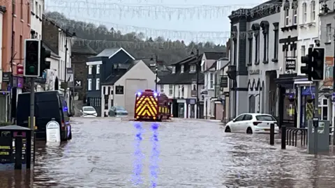 BBC Several cars and an emergency vehicle line the street with water up to their tyres